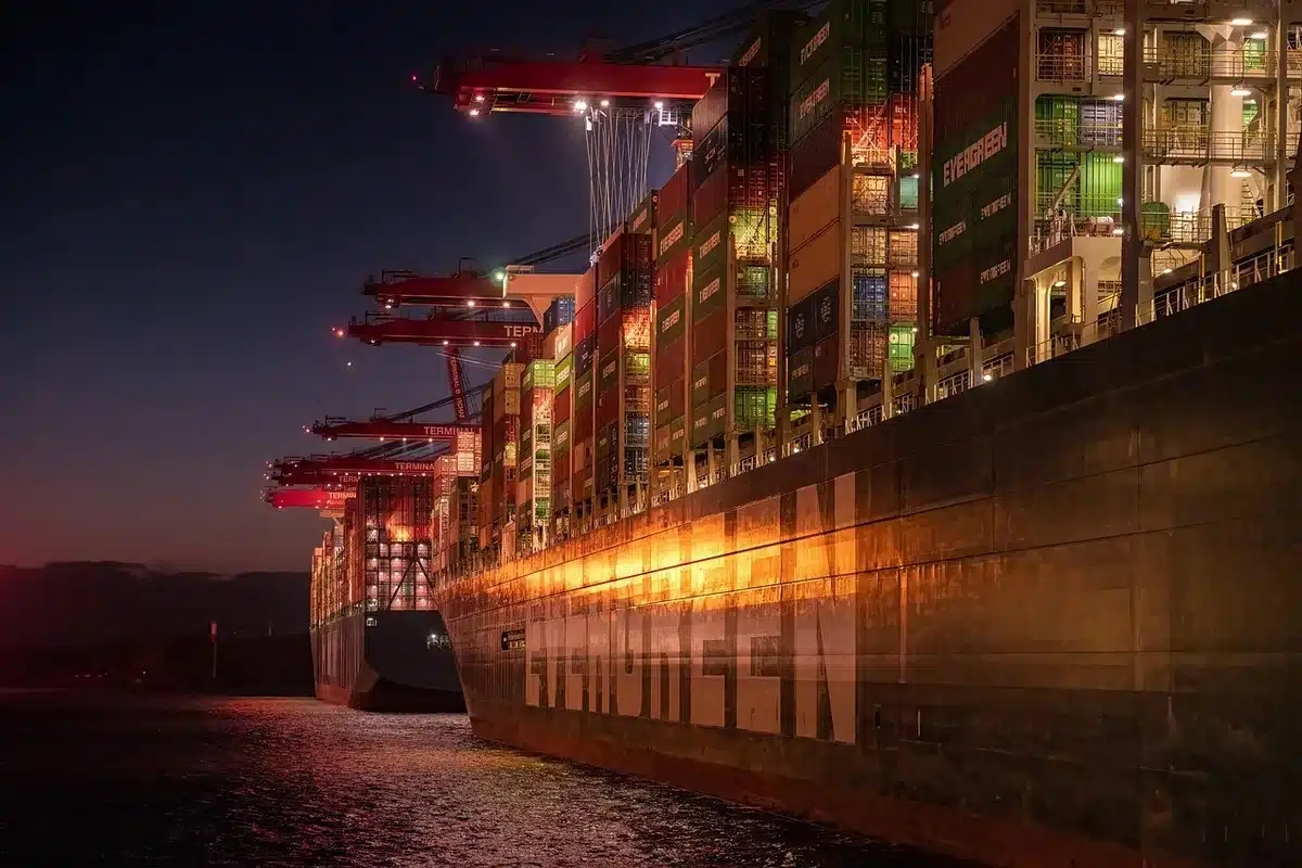 Port with cranes and stacked containers, silhouetted workers and a cargo ship in the distance